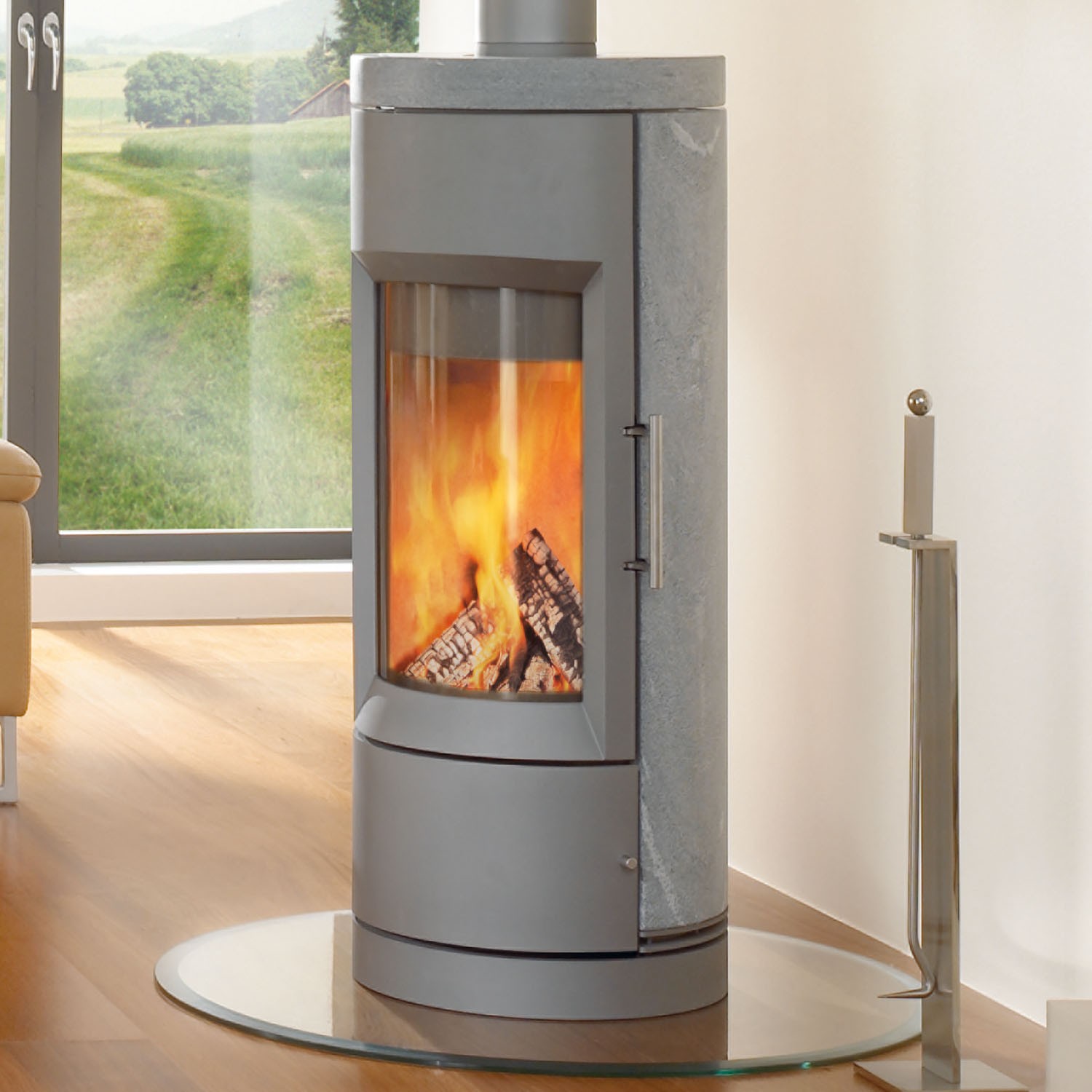 Modern round facing Bari 8170 Wood Stove with wall of windows to the left and white wall to the right with tools.