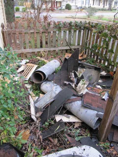 Destroyed chimney flue with roofing and other debris all over the ground. Picket fence in the background.