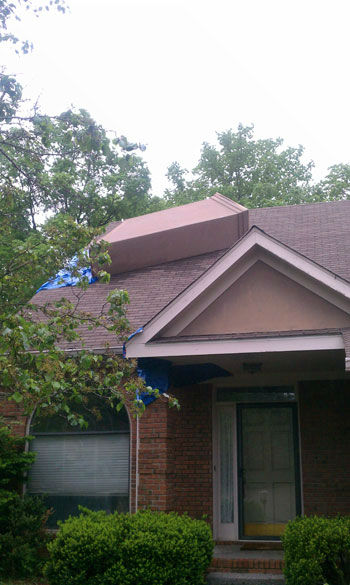 Chimney toppled over on roof with tarp covering damage. Trees in background and bushes in foreground in front of house.