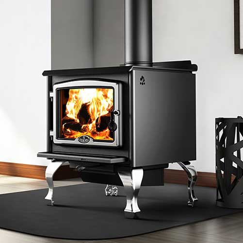 Osburn 2300 Wood Stove black with silver legs and trim around the door.  Tools to the right and white wall in the background.  There is a large flame in the stove.