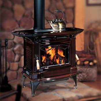 large black wood stove with four legs and kettle on top of it a fire burning in it and bricks on the wall behind