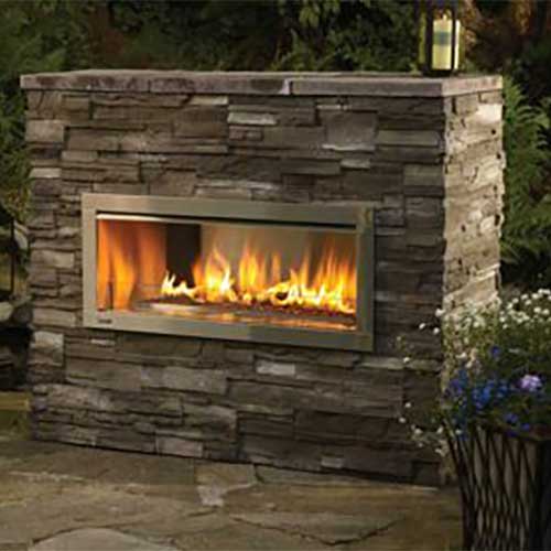 large brick outdoor fireplace with gas fireplace insert with fire buring inside it flowers are on a table to the right