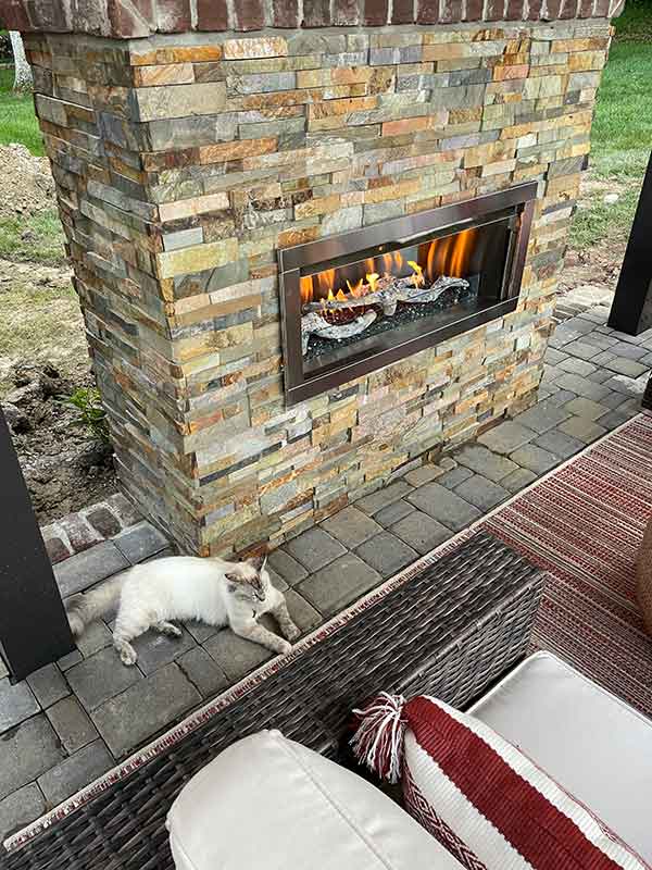 Outdoor Fireplace with nice multicolored brick on patio with pretty Himalayan kitty laying in front.  There is a red and white pillows wicker sofa in foreground.