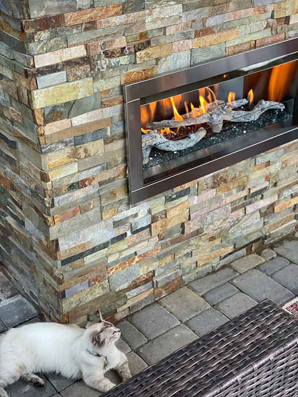 Outdoor Fireplace with stacked multi-colored stone and Himalayan kitty sitting between fireplace and wicker sofa.