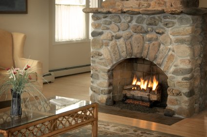 Beautiful new fireplace with river rock surround.  Cream colored chair to the left with coffee table in foreground flowers on top.  Windows in background.