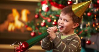 little boy with a green noisemaker in front of a christmas tree and fireplace